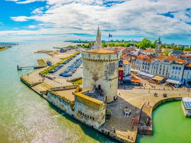 Luftaufnahme des mittelalterlichen Steinturms am Hafeneingang von La Rochelle mit Uferpromenade, parkenden Autos und historischen Gebäuden.