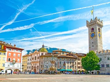 Piazza Duomo in Trient mit bunten historischen Gebäuden, Neptunbrunnen und Torre Civica Glockenturm unter blauem Himmel mit Kondensstreifen.