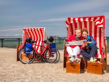 Zwei Radfahrer sitzen in einem rot-weiß gestreiften Strandkorb am Strand von Niendorf, Schleswig-Holstein. Ein Tourenrad mit blauen Taschen steht daneben.