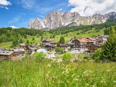Alpine village in Cortina d'Ampezzo with traditional chalets on green meadows, surrounded by dramatic Dolomite peaks under blue sky.