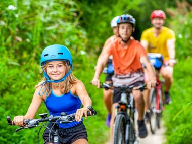 Young girl in blue helmet leads family cycling on forest path. Two cyclists follow behind through lush green vegetation in Brittany.