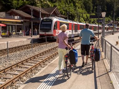 Zwei Radfahrer mit Packtaschen schieben Räder am Bahnsteig in Brienz, während ein roter Regionalzug einfährt, Berge und Gebäude im Hintergrund.