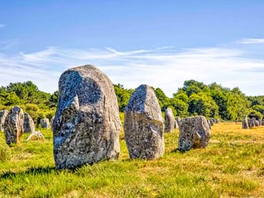 Ancient Ménec standing stones arranged in rows across a grassy field in Brittany. Large weathered megaliths stand against a blue sky with trees in the background.
