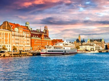 Malmö Uferpromenade mit bunten historischen Gebäuden und einem Passagierschiff am Hafen unter dramatischem Wolkenhimmel bei Sonnenuntergang.