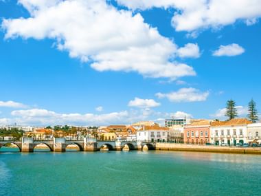 Steinbrücke mit mehreren Bögen über einen Fluss in Tavira, Algarve. Bunte historische Gebäude säumen das Ufer unter blauem Himmel.