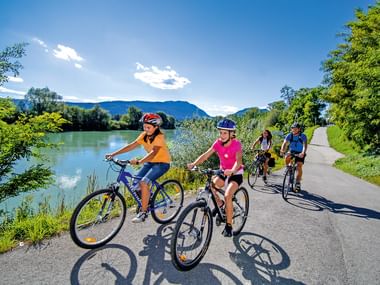 Family of four cycling on paved path beside river with mountains in background. Two children lead, followed by two adults, all wearing helmets.