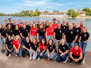 Große Teamgruppe in passenden schwarzen und roten Shirts posiert an einem Fluss mit historischen Gebäuden und Bäumen im Hintergrund.