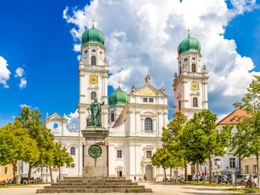 Passauer Dom mit weißer Barockfassade und zwei Türmen mit grünen Zwiebelhauben. Eine Statue steht auf dem Platz davor unter blauem Himmel.