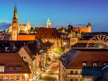 Abendansicht der Nürnberger Altstadt mit beleuchteten Kirchtürmen, Burg und Riesenrad. Historische Gebäude mit roten Dächern säumen eine Straße.