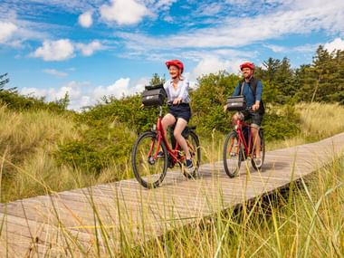 Zwei Radfahrer mit roten Helmen auf roten Rädern fahren auf einem Holzbohlenweg durch Dünengras in Föhr-Nieblum unter blauem Himmel mit weißen Wolken.