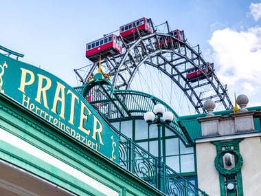 Grünes verziertes Eingangstor mit 'PRATER'-Schild in Wien, dahinter das berühmte Riesenrad mit roten Kabinen vor blauem Himmel.