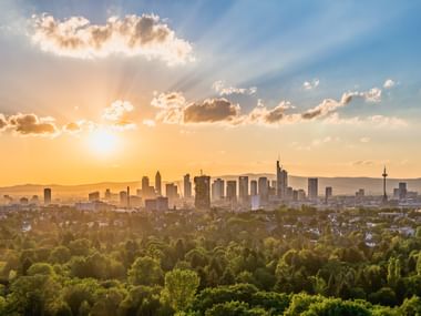 Frankfurter Skyline bei Sonnenuntergang mit Wolkenkratzern vor goldenem Himmel. Grüne Bäume und Wohngebiete im Vordergrund.
