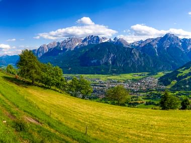 Green meadows and trees in foreground with town of Lienz in valley below. Dramatic Dolomite mountain peaks under blue sky with clouds.