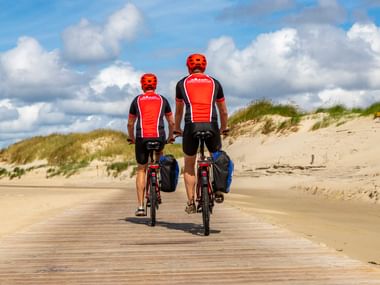 Zwei Radfahrer in roten Trikots fahren auf einem Holzbohlenweg durch Sanddünen in St. Peter-Ording unter blauem Himmel mit weißen Wolken.