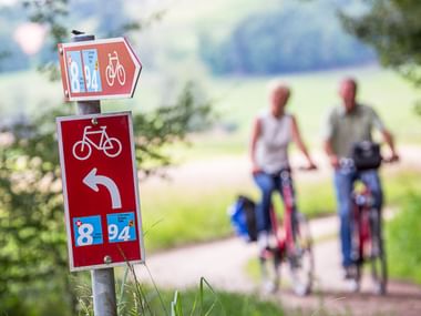 Rotes Radweg-Schild mit Route 8 und 94 und Linkspfeil am Aare-Radweg. Zwei unscharfe Radfahrer auf dem Weg im Hintergrund.