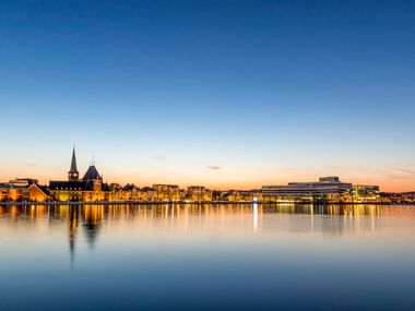 Aarhus Uferpromenade bei Sonnenuntergang mit beleuchteten Gebäuden, die sich im ruhigen Hafenwasser spiegeln. Kirchturm links, moderne Gebäude rechts.
