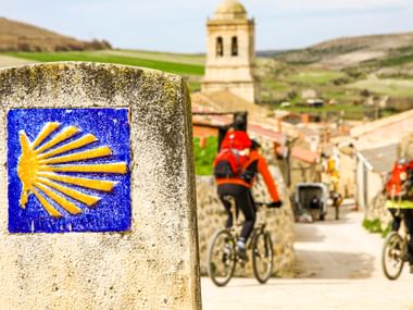 Gelbe Jakobsmuschel-Wegmarkierung auf Steinpfosten am Camino de Santiago, mit Radfahrern in einem spanischen Dorf mit Kirchturm.
