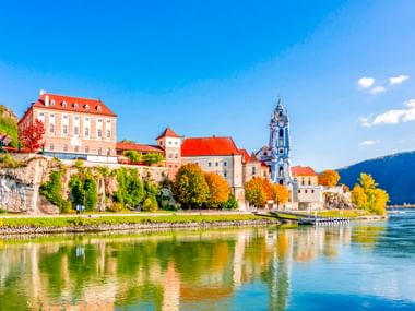 Dürnstein am Flussufer mit historischen Gebäuden und blauem Barockturm der Stiftskirche Mariä Himmelfahrt, gespiegelt in der Donau.