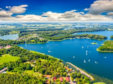Luftaufnahme der Lötzen-Seenregion in Masuren mit blauem Wasser, grünen bewaldeten Inseln, Segelbooten und vereinzelten Gebäuden unter bewölktem Himmel.