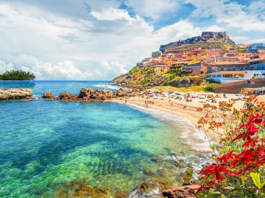 Beach bay in Castelsardo with clear turquoise water, red flowers in foreground, and hillside town with historic castle on top under blue sky.