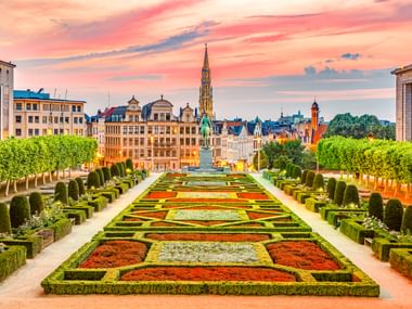 Formale Gärten am Mont des Arts in Brüssel mit bunten Blumenbeeten, geschnittenen Hecken und Stadtsilhouette unter rosa Abendhimmel.