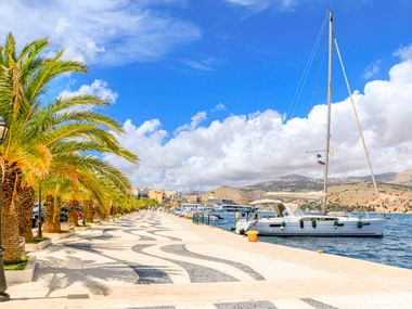 Sonnige Hafenpromenade in Argostoli mit dekorativem Wellenmuster-Pflaster, Palmen und vertäuten Segelbooten. Hügel im Hintergrund.