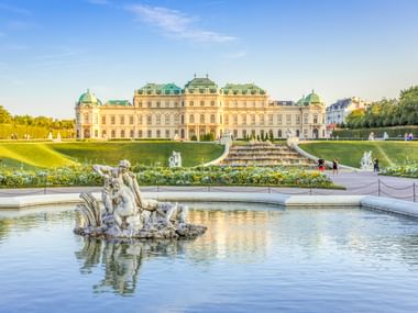 Barockes Schloss Belvedere in Wien mit türkisfarbenen Kuppeln hinter formalen Gärten. Verzierter Brunnen im Vordergrund spiegelt Sonnenlicht.