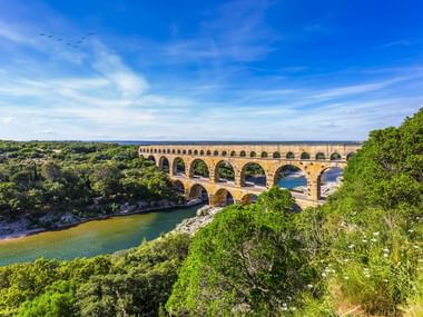 Pont du Gard, eine dreistöckige römische Aquäduktbrücke bei Remoulins, die einen Fluss überspannt, umgeben von üppiger grüner Vegetation unter blauem Himmel.