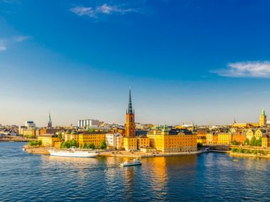 Panoramablick auf Stockholms Gamla Stan mit bunten historischen Gebäuden am Wasser, Kirchturm und Booten auf dem Wasser unter blauem Himmel.