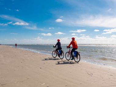 Zwei Radfahrer fahren am Sandstrand der Insel Rømø entlang, das Meer zu ihrer Rechten. Blauer Himmel mit weißen Wolken über dem breiten Strand.