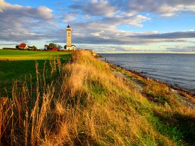Weißer Helnæs Leuchtturm auf grasbewachsener Küstenklippe mit Blick auf die Ostsee. Rotes Bauernhaus und grüne Felder unter bewölktem Himmel.