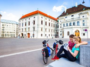 Paar mit Tourenrädern rastet auf Steinbank am Hofburg-Platz in Wien, mit historischen weißen Palastgebäuden mit roten Dächern.