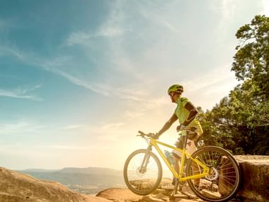 Cyclist in green jersey and helmet standing with yellow mountain bike on rocky viewpoint overlooking valley and mountains under blue sky.