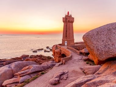 Phare de Ploumanac'h lighthouse on pink granite rocks at sunset. The stone tower overlooks the Atlantic Ocean with warm orange sky.