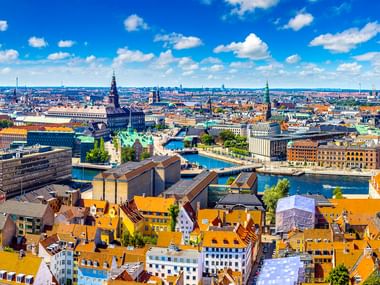 Panoramablick auf Kopenhagen mit orangefarbenen Ziegeldächern, Kirchtürmen, Wasserstraßen und modernen Gebäuden unter blauem Himmel.