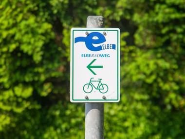 Weißes Wegweiserschild für den Elberadweg mit blauem Logo, grünem Pfeil nach links und Fahrradsymbol, an Metallpfosten montiert.