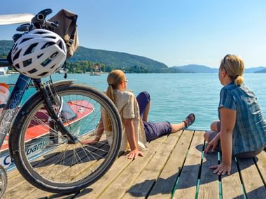 Zwei Radfahrer sitzen auf einem Holzsteg im Hafen Velden mit einem Fahrrad im Vordergrund. Berge und ruhiges Seewasser im Hintergrund.
