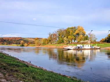 Fähre überquert die Weser bei Polle mit herbstlichem Laub an den Ufern. Grüne Grasflächen führen zum ruhigen Wasser unter bewölktem Himmel.