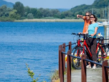 Zwei Radfahrer mit roten Fahrrädern auf einem Uferweg am Überlinger See, Bodensee. Blaues Wasser, Segelboote und grüne Hügel im Hintergrund.