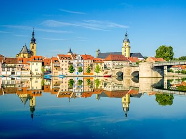Historische Steinbrücke über den Main in Kitzingen mit bunten Häusern und Kirchtürmen, die sich im ruhigen Wasser unter blauem Himmel spiegeln.
