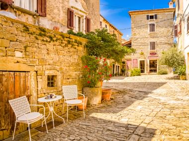 Sonnige Kopfsteinpflasterstraße in Grožnjan mit Steingebäuden, weißen Caféstühlen, Topfpflanzen und blauem Himmel. Historische kroatische Hügelstadt.