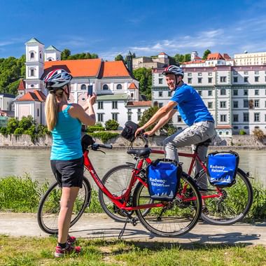 Zwei Radfahrer mit Helmen und Packtaschen pausieren an einem Fluss in Passau. Historische Gebäude mit orangen Dächern und Kirchturm am Ufer.