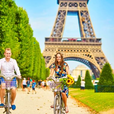 Mann und Frau radeln auf baumgesäumtem Weg mit Eiffelturm im Hintergrund. Frau trägt Sonnenblumen im Fahrradkorb an sonnigem Tag in Paris.