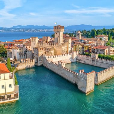 Luftaufnahme der Scaligerburg in Sirmione am Gardasee mit befestigten Mauern im türkisfarbenen Wasser, umgeben von rotgedeckten Gebäuden.