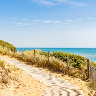 Wooden boardwalk winding through sandy dunes with beach grass and wooden fencing at Pointe du Payre, Jard-sur-Mer, leading to turquoise ocean.