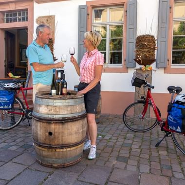 Zwei Radfahrerinnen probieren Rotwein an Holzweinfässern vor einem traditionellen weißen Gebäude in Maikammer, Tourenräder in der Nähe.
