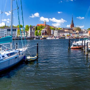 Flensburger Hafen mit Segelbooten an Holzstegen. Historische Gebäude und Kirchtürme säumen die Uferpromenade unter blauem Himmel mit Wolken.