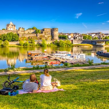 Familie beim Picknick am Ufer der Maine mit Festungsmauern und Türmen des Schlosses Angers im Hintergrund. Boote am Flussufer unter blauem Himmel.