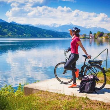 Radfahrerin in pinkem Trikot mit Helm steht mit Fahrrad auf Plattform am Seeufer in Österreich, Blick auf Berge und See unter blauem Himmel.
