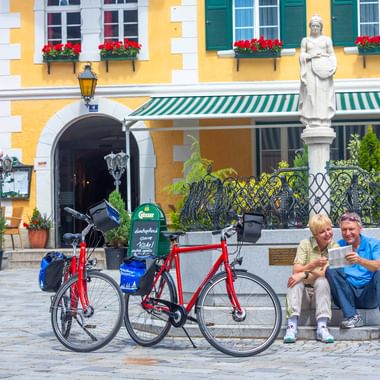 Two cyclists with touring bikes rest at a fountain with statue in front of a yellow building with red geraniums in Oberdrauburg.